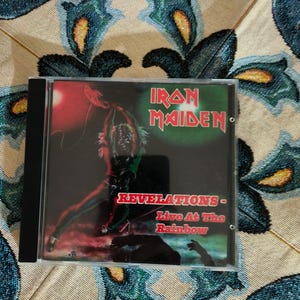 Op de afbeelding: Een cd-hoesje met het Iron Maiden-album "Revelations - Live At The Rainbow". De donkere cover toont een actiescène met het logo van de band in groene en rode tekst. De hoes zit in een jewelcase.
