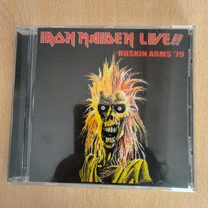 Könnte beinhalten: Eine CD-Hülle mit dem Album "Iron Maiden Live!! Ruskin Arms '79". Das Cover zeigt eine skelettartige Figur mit gelben und orangenen Flammen als Haare und einem gelben Gewand vor schwarzem Hintergrund. Der Bandname ist rot.