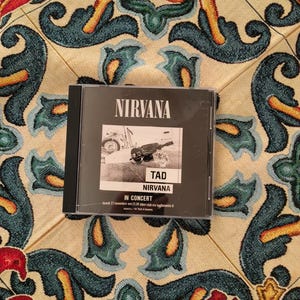 May include: A black and white Nirvana concert CD case featuring the text NIRVANA, TAD, and IN CONCERT. The cover art displays a monochrome photograph of a person with a guitar, with additional event details printed in small text at the bottom.