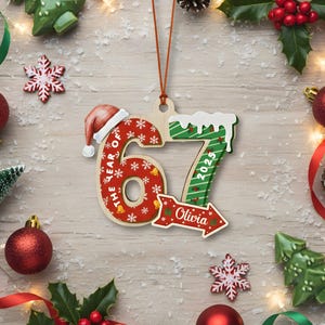 May include: Wooden Christmas ornament shaped like the number 67 with a Santa hat on top. The number 6 is red with white snowflakes and the text "THE YEAR OF" and the number 7 is green with white snow and the year "2023". An arrow with the name "Olivia" is also included.