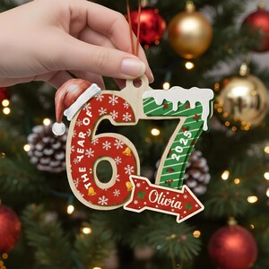 May include: Wooden Christmas ornament shaped like the number 67 with a Santa hat on the 6. The 6 is red with snowflakes and bells. The 7 is green with white snow and the year 2025. A red arrow with the name "Olivia" is attached.