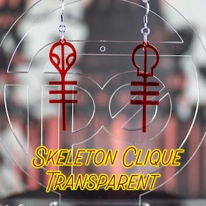 May include: A pair of red acrylic earrings with a skeleton clique logo. The earrings are hanging from a clear acrylic display stand. The text "SKELETON CLIQUE TRANSPARENT" is printed on the stand.