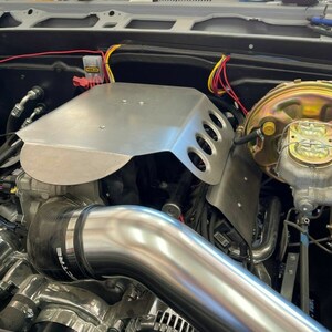 May include: Close-up of a car engine with a silver metal cover, a silver exhaust pipe, and a black and silver air filter. The engine is surrounded by wires and hoses.