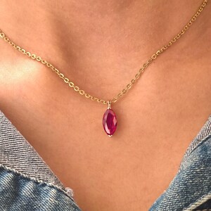 May include: A gold-colored chain necklace with a marquise-cut, ruby-red pendant. The pendant is suspended from a delicate chain, creating a simple yet elegant design. The necklace is worn against a denim background.