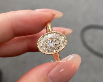 May include: A gold ring with an oval-cut diamond. The diamond is set in a bezel setting with a beaded edge. The ring is held by a person with manicured nails. The text "ParryGoldJewels" is visible on the ring.
