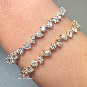 Puede incluir: Dos pulseras de diamantes, una dorada y otra plateada, se muestran en una muñeca. Las pulseras presentan una variedad de cortes de diamantes, incluyendo formas redondas, de pera y esmeralda. Las piedras preciosas son transparentes.