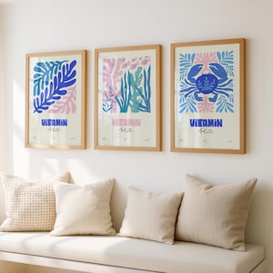 May include: Three framed art prints with a beach theme. Each print features a different design in blue, pink, and green, with the words "Vitamin Sea" at the bottom. The frames are a light wood color, and the prints are displayed above a white bench with pillows.