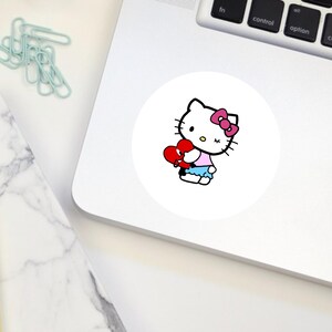 Puede incluir: Un personaje blanco de Hello Kitty con un lazo rosa sosteniendo un coraz&oacute;n rojo con una venda blanca. El personaje lleva una camisa rosa y una falda azul.