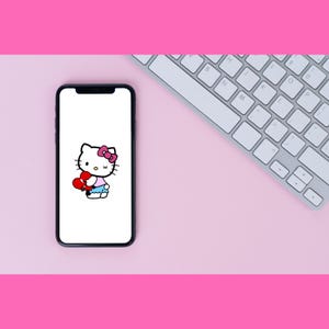 Puede incluir: Un tel&eacute;fono inteligente blanco con una pantalla negra que muestra una imagen de dibujos animados de Hello Kitty sosteniendo un coraz&oacute;n rojo. El fondo es rosa.