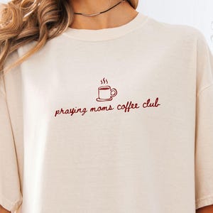 Op de afbeelding: Ivoor kleurig T-shirt met de tekst "praying moms coffee club" in bordeauxrode letters, samen met een kleine koffiekopje afbeelding. Het shirt heeft een relaxte pasvorm en korte mouwen. De kleur is ivoor.