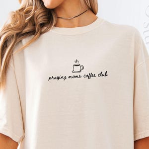 Op de afbeelding: Ivoor kleurig t-shirt met de tekst "praying moms coffee club" in zwart schrift. Een kleine koffiekopje afbeelding staat boven de tekst. Het shirt heeft een relaxte pasvorm en korte mouwen. De kleur is ivoor.