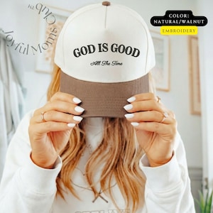 May include: A baseball cap with a white crown and a brown brim. The text "GOD IS GOOD All The Time" is embroidered in black on the front. The cap is labeled "NATURAL/WALNUT" and "EMBROIDERY".