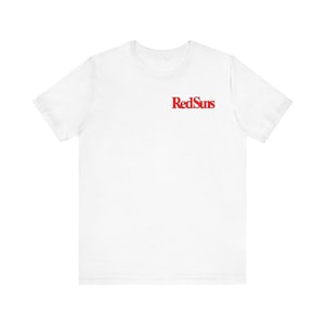 May include: A white short-sleeved t-shirt featuring the text RedSuns printed in a bold red serif font on the upper left chest area. The garment has a classic crew neckline and a standard fit, providing a clean and simple aesthetic for casual wear.