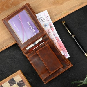 May include: A brown leather wallet with a clear plastic window for a card. The wallet has multiple card slots, a billfold, and a small pocket. The wallet is open and has a card with the text "Heathrow Rewards" and a few banknotes inside.