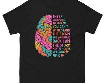 Può includere: T-shirt nero con grafica a cervello colorata e la scritta "THEY WHISPERED TO HER YOU CAN'T WITH STAND THE STORM SHE WHISPERED BACK I AM THE STORM MENTAL HEALTH WARRIOR". Girocollo classico.