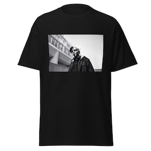 May include: Black t-shirt with a black and white image of a person wearing a mask and a jacket. The image on the shirt is a photograph of a person standing in front of a building. The t-shirt is made of a soft material.