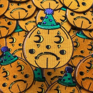 May include: Embroidered orange patch featuring a sad-faced pumpkin wearing a green party hat with purple trim. The pumpkin has crescent moon eyes and black dots. The patches are scattered across the image.