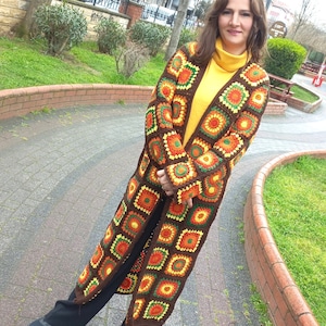 May include: A long, open-front crochet cardigan in a patchwork design. The cardigan features square motifs in shades of brown, orange, yellow, and green. The person is wearing a yellow turtleneck and black trousers. The cardigan is knee-length.