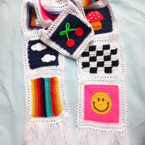 Puede incluir: Una larga bufanda de crochet blanca con cuadrados coloridos que representan un arcoíris, un patrón de cuadros, una cara sonriente, un hongo, una cereza y nubes. La bufanda tiene flecos blancos en ambos extremos.