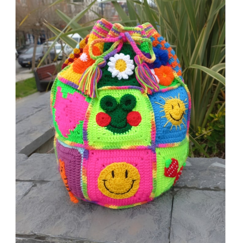 Op de afbeelding: Een kleurrijke gehaakte trekkoordtas met verschillende patches, waaronder een smiley, een aardbei, een bloem en een kikker. De tas is gemaakt met felle kleuren zoals roze, groen, geel en blauw.