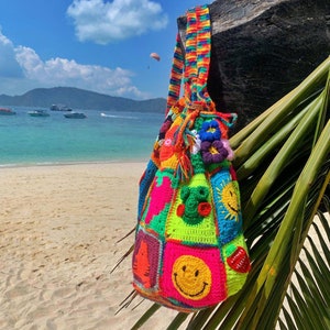 Op de afbeelding: Een kleurrijke gehaakte tas met verschillende smiley-gezichten, dieren en andere ontwerpen. De tas heeft een lange regenboogband en hangt aan een houten paal op een strand.