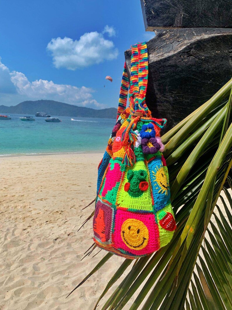 Op de afbeelding: Een kleurrijke gehaakte tas met verschillende smiley-gezichten, dieren en andere ontwerpen. De tas heeft een lange regenboogband en hangt aan een houten paal op een strand.