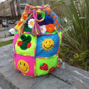 Op de afbeelding: Een kleurrijke gehaakte trekkoordtas met verschillende ontwerpen, waaronder smiley-gezichten, bloemen en aardbeien. De tas is gemaakt met felle kleuren zoals roze, groen, blauw en geel.