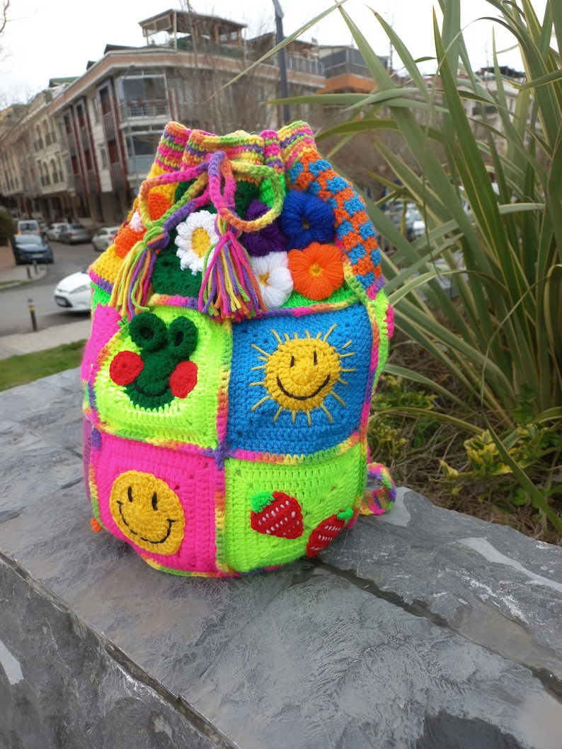 Op de afbeelding: Een kleurrijke gehaakte trekkoordtas met verschillende ontwerpen, waaronder smiley-gezichten, bloemen en aardbeien. De tas is gemaakt met felle kleuren zoals roze, groen, blauw en geel.