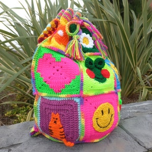 Op de afbeelding: Een kleurrijke gehaakte tas met verschillende ontwerpen, waaronder een hart, een smiley, een kat en een bloem. De tas is gemaakt van verschillende gekleurde garens en heeft een trekkoord.