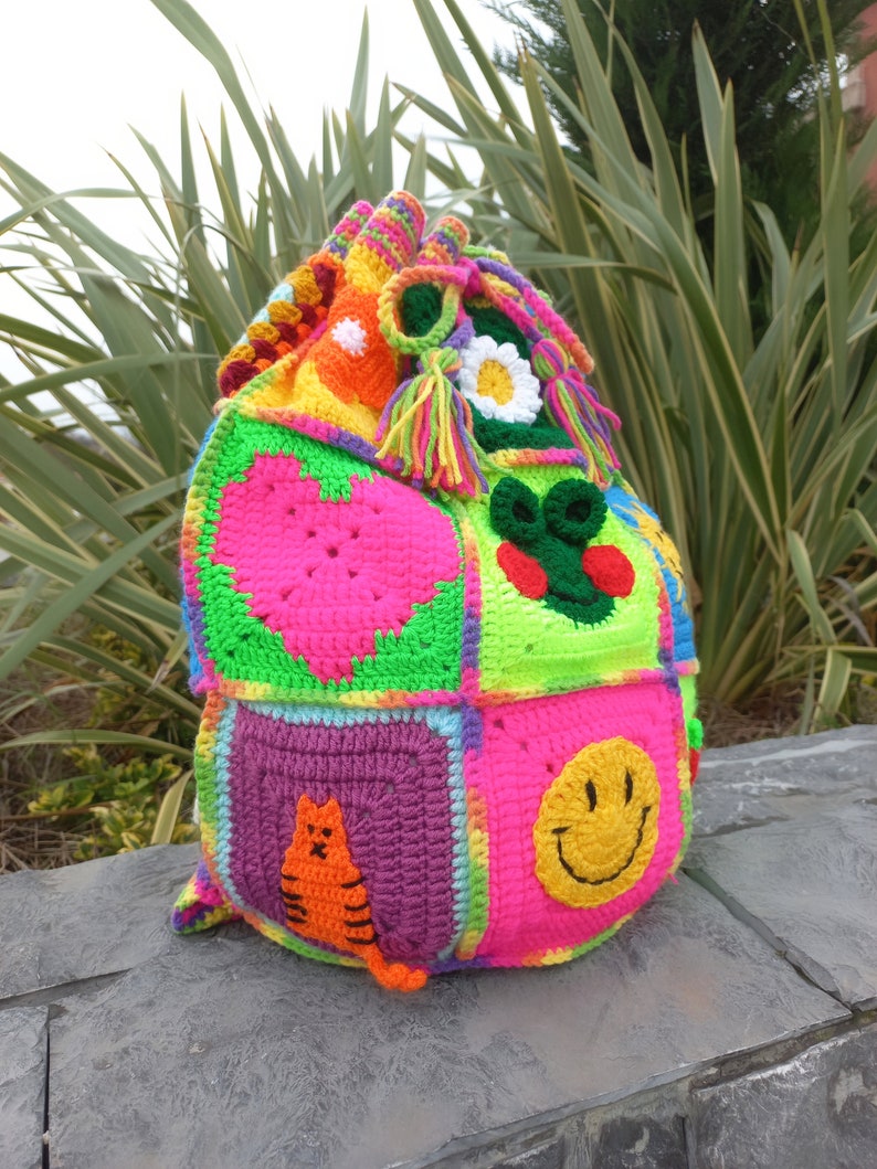 Op de afbeelding: Een kleurrijke gehaakte tas met verschillende ontwerpen, waaronder een hart, een smiley, een kat en een bloem. De tas is gemaakt van verschillende gekleurde garens en heeft een trekkoord.