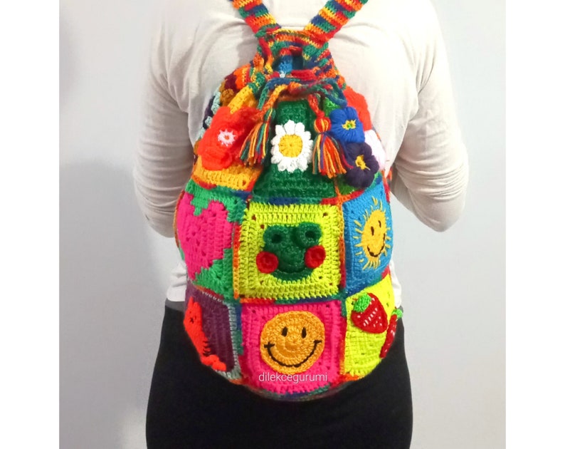Op de afbeelding: Een kleurrijke gehaakte rugzak met een trekkoord. De rugzak is gemaakt van vierkanten met verschillende ontwerpen, waaronder een kikker, een zon, een aardbei en een smiley. De rugzak heeft een regenboogband en een kwast.