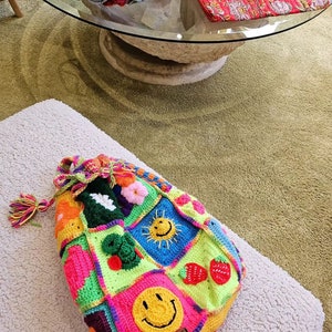 Op de afbeelding: Een kleurrijke gehaakte tote bag met verschillende ontwerpen, waaronder een smiley, een zon en aardbeien. De tas heeft een lange, kleurrijke kwast.