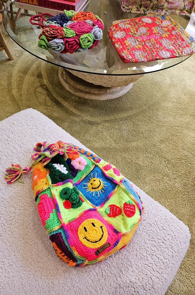 Op de afbeelding: Een kleurrijke gehaakte tote bag met verschillende ontwerpen, waaronder een smiley, een zon en aardbeien. De tas heeft een lange, kleurrijke kwast.