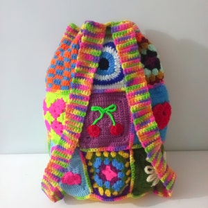 Op de afbeelding: Een kleurrijke gehaakte rugzak met verschillende patronen, waaronder een regenboogstreep, een boze oog en kersen. De rugzak is gemaakt van verschillende gekleurde garen en heeft een unieke, handgemaakte uitstraling.