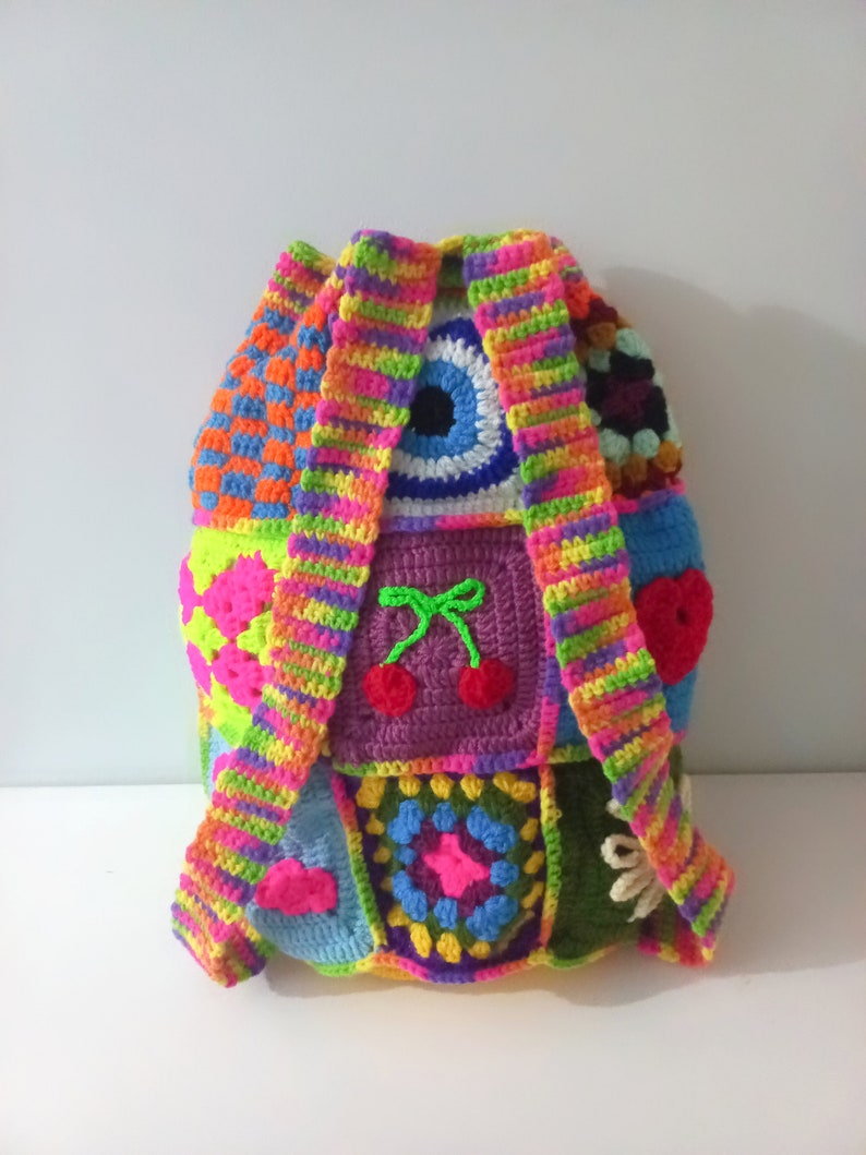 Op de afbeelding: Een kleurrijke gehaakte rugzak met verschillende patronen, waaronder een regenboogstreep, een boze oog en kersen. De rugzak is gemaakt van verschillende gekleurde garen en heeft een unieke, handgemaakte uitstraling.