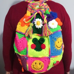 Op de afbeelding: Een kleurrijke gehaakte rugzak met verschillende patches, waaronder een hart, een zon, een smiley en een bloem. De rugzak heeft een felgroene, roze, blauwe en gele kleurstelling.