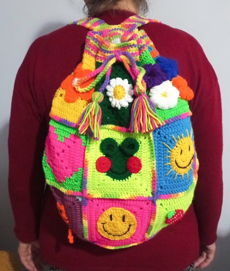 Op de afbeelding: Een kleurrijke gehaakte rugzak met verschillende patches, waaronder een hart, een zon, een smiley en een bloem. De rugzak heeft een felgroene, roze, blauwe en gele kleurstelling.