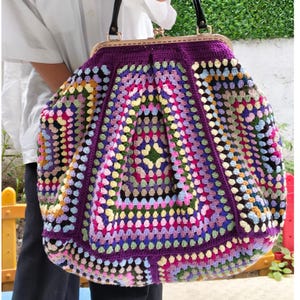 May include: A large, handmade crochet bag with a purple frame and handles. The bag features a colorful patchwork design with squares in various colors, including pink, blue, yellow, and green. The bag is held by a person wearing a white shirt.