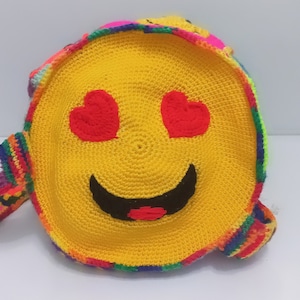 Op de afbeelding: Een gele gehaakte tas met een smiley-gezicht en hartjesogen. De tas heeft een regenboogkleurige rand en een lange band.