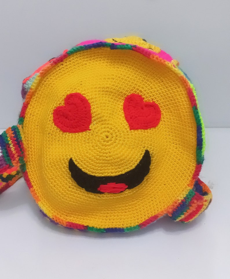 Op de afbeelding: Een gele gehaakte tas met een smiley-gezicht en hartjesogen. De tas heeft een regenboogkleurige rand en een lange band.