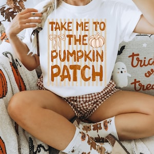 Pode incluir: Uma t-shirt branca de manga curta com um gráfico de riscas verticais bege e o texto laranja "TAKE ME TO THE PUMPKIN PATCH". O design inclui uma ilustração linear de um laço e uma abóbora sobre um fundo casual de temática outonal.