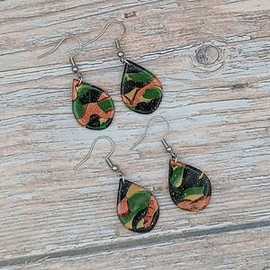 Peut inclure: Deux paires de boucles d'oreilles pendantes en forme de goutte d'eau avec un motif camouflage dans des tons de noir, vert et cuivre. Chaque boucle est suspendue à un crochet en métal argenté. La surface des pendentifs a une finition pailletée.