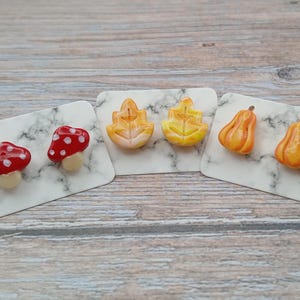 May include: Three pairs of stud earrings displayed on white marble-patterned cards. One pair features red mushrooms with white spots, one pair shows yellow and orange leaves, and the third pair consists of orange pumpkins with green stems.