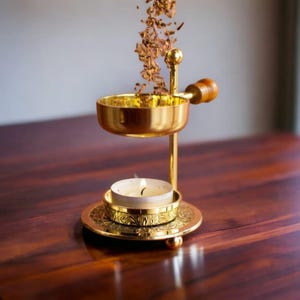 May include: A gold metal incense burner with a round top and a wooden handle. The burner is on a wooden table and has a small white candle underneath it. Incense is falling from the top of the burner.