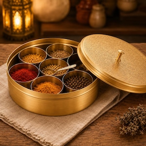 Puede incluir: Una caja de especias dorada con tapa decorativa, que contiene varias especias en compartimentos individuales. La caja está sobre una tela beige, con una superficie de madera en el fondo. Las especias son rojas, amarillas y marrones.