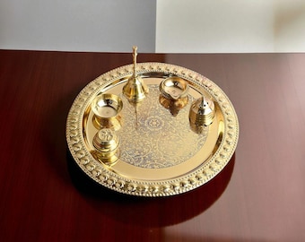 May include: A gold-coloured puja thali set with a floral design. The set includes a bell, small bowls, a lamp, and a container. The thali is round with a decorative beaded edge. The items are arranged on a dark wooden surface.