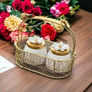 Op de afbeelding: Twee witte keramische potten met gouden deksels en witte bloemenaccenten worden gepresenteerd in een gouden draadmand met een handvat. De mand staat op een houten oppervlak.