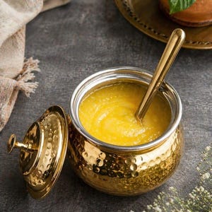 May include: A gold-colored, hammered-metal serving bowl with a lid and a spoon. The bowl contains a yellow, creamy substance. The lid is off to the side. The bowl is on a gray surface.