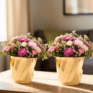 May include: Two gold vases with a spiral design, each holding a floral arrangement of pink, white, and green flowers. The flowers include chrysanthemums and roses, creating a colourful display.