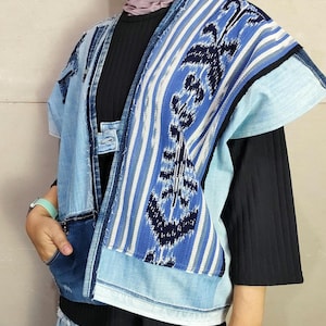 May include: A blue denim vest with a patterned fabric panel on the front. The vest has a collar and short sleeves. The fabric panel is a mix of blue, white and black stripes with a geometric design.